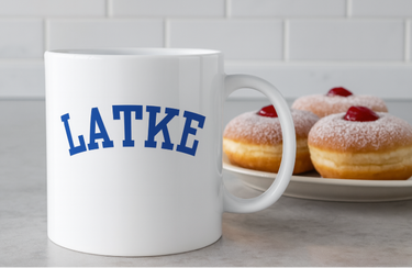 White mug with 'LATKE' in blue text, placed on a surface with donuts in the background.