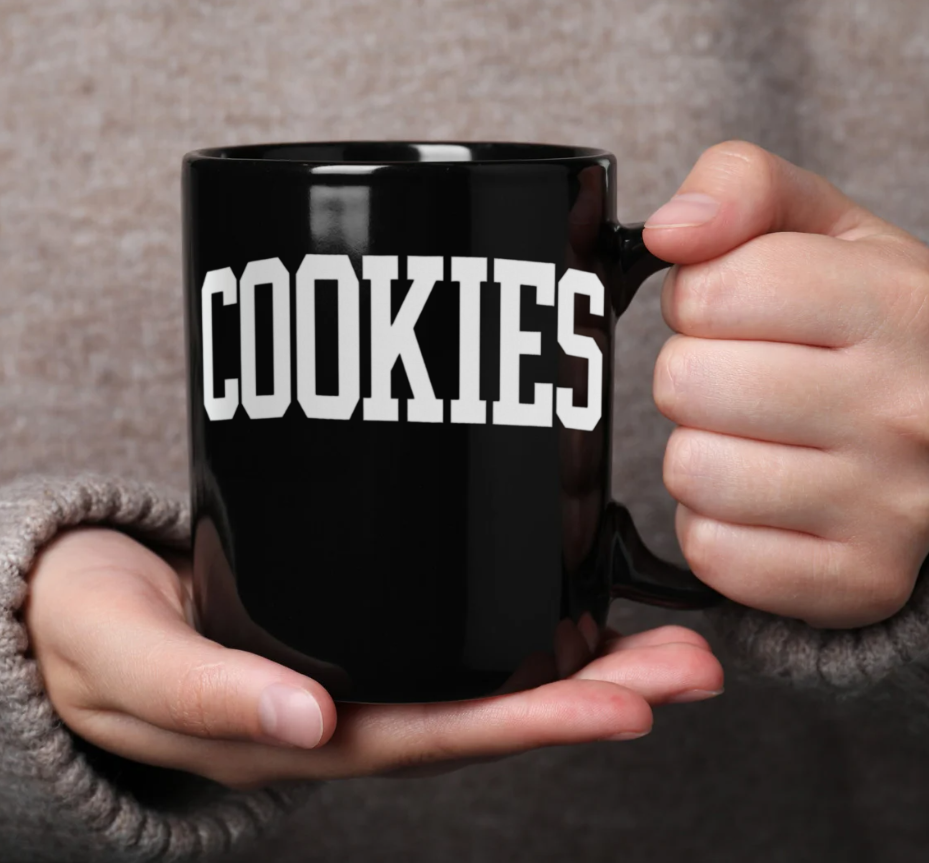 Black mug with 'COOKIES' text held by a person against a neutral background