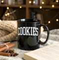 Black mug with 'COOKIES' text on a cozy setting with cinnamon sticks and an open book.