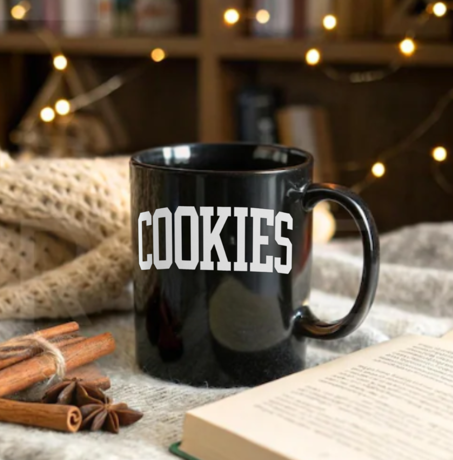 Black mug with 'COOKIES' text on a cozy setting with cinnamon sticks and an open book.
