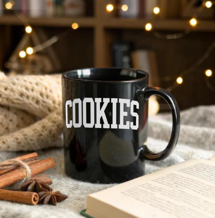 Black mug with 'COOKIES' text on a cozy setting with cinnamon sticks and an open book.