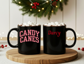 Two black mugs with 'Candy Canes' and 'Darcy' text, topped with marshmallows, on a wooden coaster.