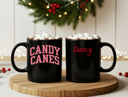 Two black mugs with 'Candy Canes' and 'Darcy' text, topped with marshmallows, on a wooden coaster.