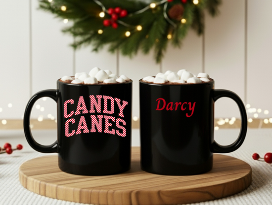 Two black mugs with 'Candy Canes' and 'Darcy' text, topped with marshmallows, on a wooden coaster.