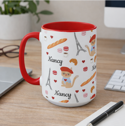 White mug with red interior and handle, featuring Paris-themed illustrations. a beret wearing cat and the name 'Nancy', on a desk.