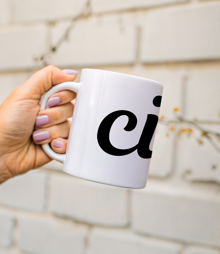 Hand holding a white mug with 'ciao" printed on it against a light brick wall.