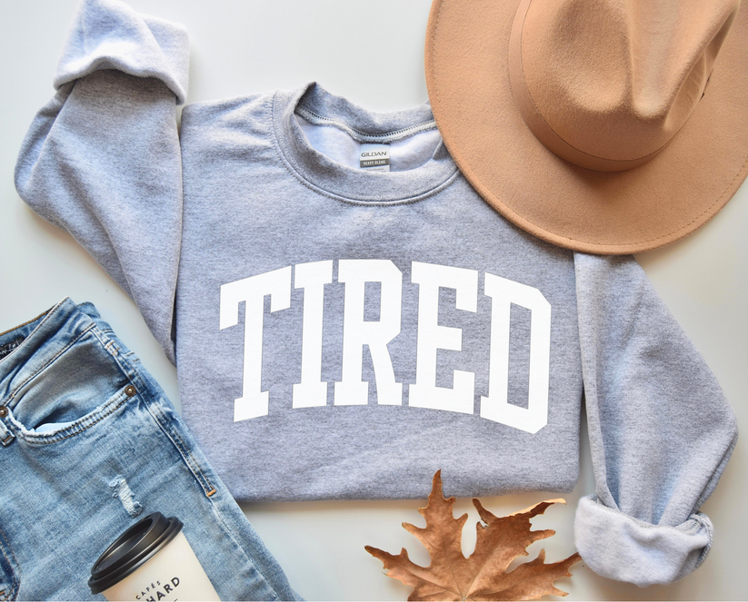 Gray sweatshirt with 'TIRED' text, denim jeans, tan hat, and coffee cup on a light surface.