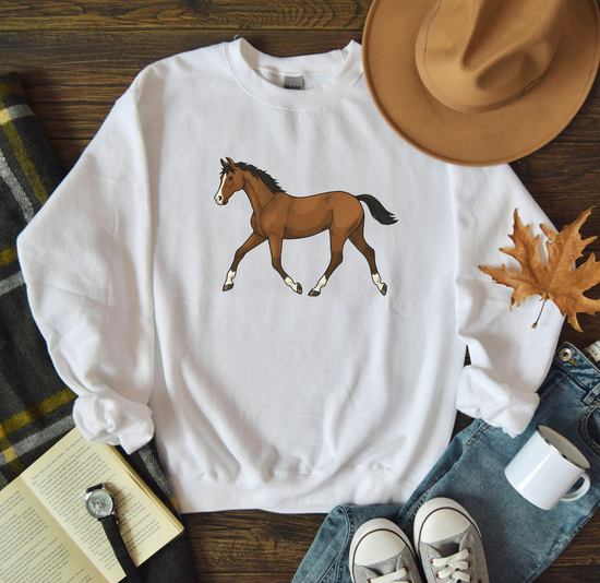 White sweatshirt with a horse graphic on a wooden surface with a hat, mug, and book.