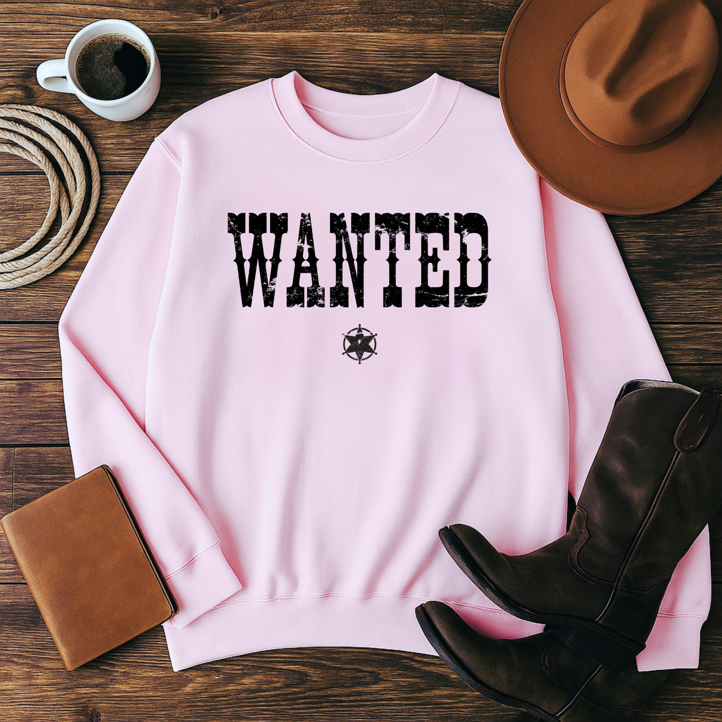 Pink sweatshirt with 'WANTED' text on a wooden surface with cowboy hat, boots, and coffee.