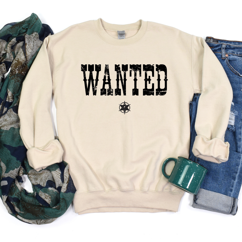 Beige sweatshirt with 'WANTED' print, surrounded by camouflage fabric, jeans, and a green mug.
