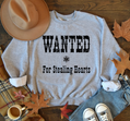Gray sweatshirt with 'WANTED For Stealing Hearts' text on a wooden surface with autumn leaves and a hat.