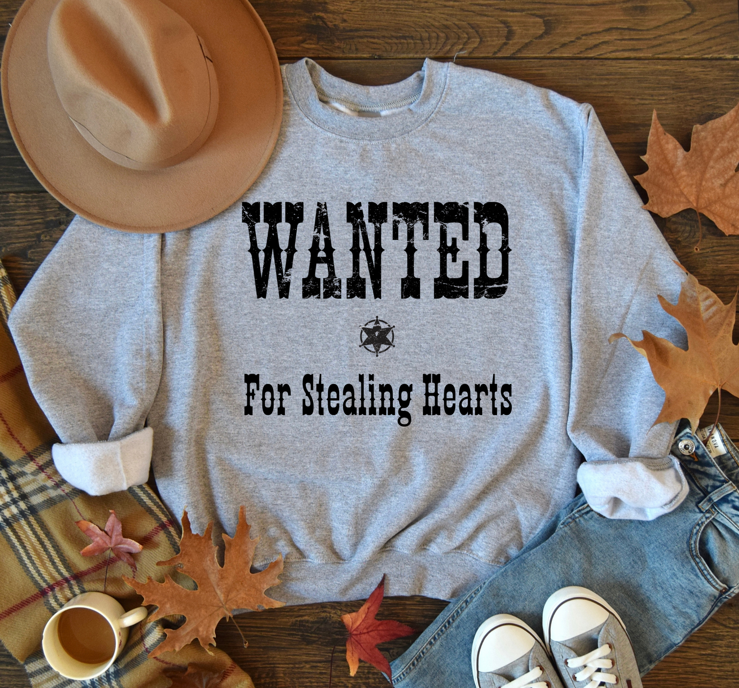 Gray sweatshirt with 'WANTED For Stealing Hearts' text on a wooden surface with autumn leaves and a hat.