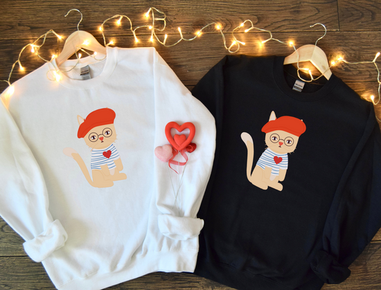 White and black sweatshirts with cartoon cat design on a wooden surface with string lights.