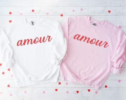 Two sweatshirts, one white and one pink, with 'amour' text on a wooden surface with heart-shaped confetti.