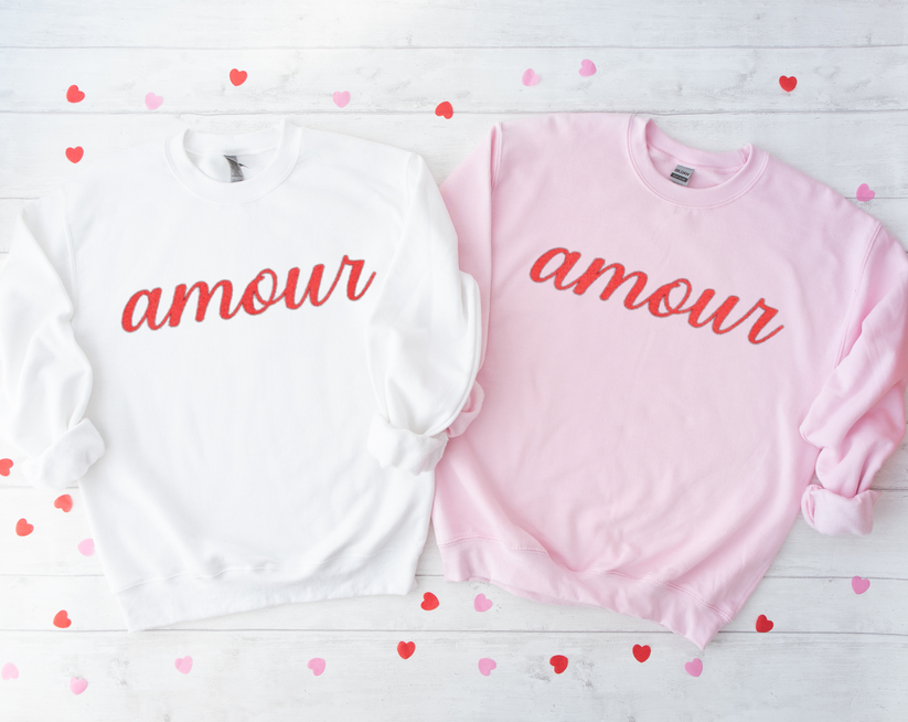Two sweatshirts, one white and one pink, with 'amour' text on a wooden surface with heart-shaped confetti.