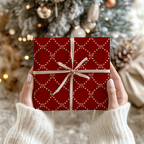 Red and gold art deco elegant patterned gift wrap on a  gift box with white and gold ribbon held by hands in front of a Christmas tree.